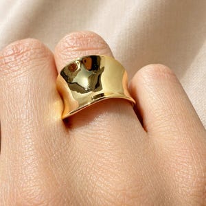 May include: A gold-colored ring with a wide, curved band. The ring has a smooth, polished surface that reflects light, creating highlights and shadows. The ring is worn on a finger.