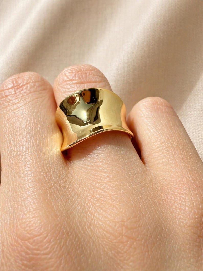 May include: A gold-colored ring with a wide, curved band. The ring has a smooth, polished surface that reflects light, creating highlights and shadows. The ring is worn on a finger.