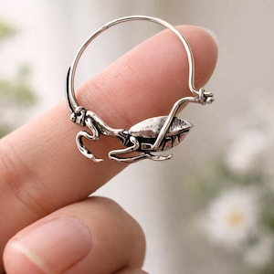 Puede incluir: Un pendiente plateado con un diseño detallado de insecto. El pendiente tiene un aro circular con una figura de insecto adjunta, que muestra detalles intrincados. El insecto parece ser un escarabajo o criatura similar, con un cuerpo texturizado y patas delicadas.