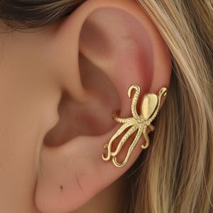 Könnte beinhalten: Goldfarbene Kraken-Ohrklemme. Die Ohrklemme zeigt ein detailliertes Kraken-Design mit acht Tentakeln und einem Kopf. Der Schmuck wird am Ohr getragen und besteht aus einem glänzenden, metallischen Material.