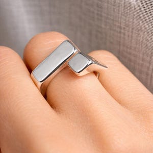 Könnte beinhalten: Ein silberner Ring mit modernem Design. Der Ring hat zwei rechteckige Formen, eine lange und eine quadratische, die in einem Winkel verbunden sind. Der Ring wird am Finger getragen.