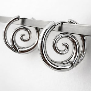 Könnte beinhalten: Ein Paar glänzende, silberfarbene Spiralohrringe. Die Ohrringe sind kreisförmig mit einem Spiraldesign in der Mitte. Sie hängen an einem silbernen Stab vor einem weißen Hintergrund. Die Ohrringe haben eine polierte, reflektierende Oberfläche.