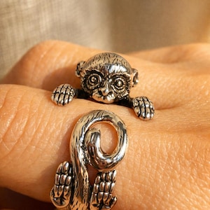 Könnte beinhalten: Ein silberner Affenring. Der Ring hat ein detailliertes Affen-Design, wobei Kopf und Arme des Affen den Finger zu umfassen scheinen. Der Schwanz des Affen bildet das Band des Rings. Der Ring ist aus glänzendem Metall gefertigt.