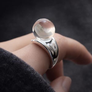 Könnte beinhalten: Ein silberner Ring mit einem klaren, kugelförmigen Edelstein. Das Band des Rings hat ein glattes, geschwungenes Design, und der Edelstein scheint über dem Finger zu schweben. Der Ring wird vor einem dunklen Hintergrund gezeigt.