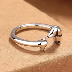 Könnte beinhalten: Ein silberfarbener offener Ring mit einem geformten Dackel-Design. Das Ringband bildet den Körper des Hundes, wobei der Kopf und die Vorderpfoten an den offenen Enden des Rings positioniert sind. Das Metall hat ein poliertes Finish.
