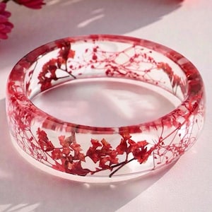 May include: Clear resin bangle bracelet with embedded red flowers. The transparent bracelet showcases the delicate floral arrangement, creating a unique and eye-catching accessory. The bangle is a circular shape.