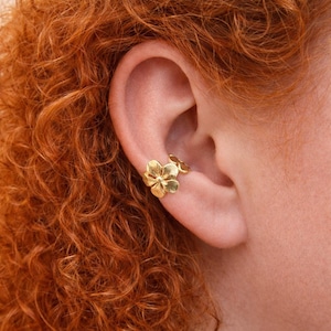 Puede incluir: Un brazalete de oreja dorado con un diseño floral. El brazalete se envuelve alrededor de la oreja, con dos acentos de flores. La joya se lleva en la oreja de una persona con el pelo rizado rojo.