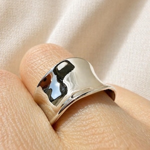 Könnte beinhalten: Nahaufnahme eines polierten Silberrings. Der Ring hat ein breites, geschwungenes Design mit einer glatten, reflektierenden Oberfläche. Der Ring befindet sich an einem Finger, und der Hintergrund ist eine weiche, neutrale Farbe.