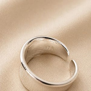 May include: A close-up of a silver ring with a smooth, polished surface. The ring has a modern, open design with a slight gap. The interior of the ring is stamped with "925". The ring is set against a neutral background.