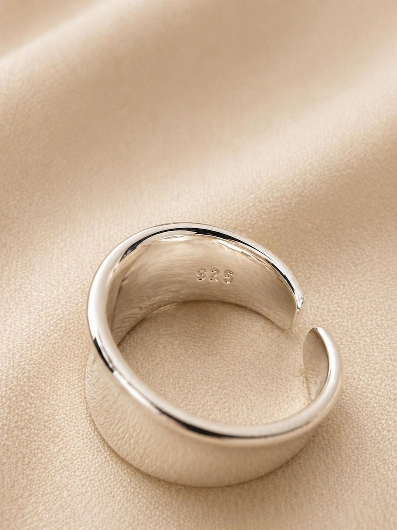 May include: A close-up of a silver ring with a smooth, polished surface. The ring has a modern, open design with a slight gap. The interior of the ring is stamped with "925". The ring is set against a neutral background.