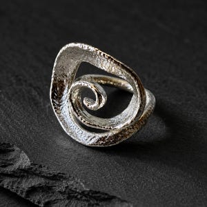 Könnte beinhalten: Ein silberfarbener Ring mit einem markanten, offen gestalteten Spiraldesign. Die Metalloberfläche weist eine ausgeprägte, raue und texturierte Struktur auf, wobei dunkle Oxidation in den Vertiefungen für ein kontrastreiches Erscheinungsbild sorgt.