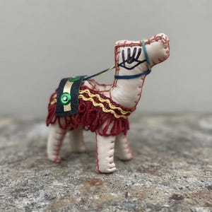 May include: Handmade camel figurine with cream-colored body, red stitching, and decorative saddle. The saddle is black and green with a gold trim and a green button. The camel has a blue and yellow harness and a decorative eye design.