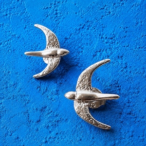 May include: Two silver-toned metal swallow bird pins featuring a textured wing surface and a smooth, polished body. Each bird is depicted in a flying position with wings extended, showcasing a metallic finish and detailed wing patterns.