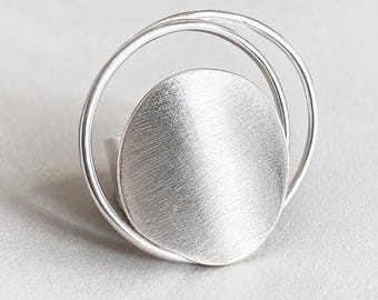 May include: A silver ring featuring a large, circular centrepiece with a brushed, textured finish. The centrepiece is framed by two thin, smooth, polished silver wire circles that extend outward, creating a layered, open, and geometric design.