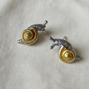 以下が含まれることがあります： カタツムリ型のスタッドピアス。ピアスは、テクスチャード加工が施されたシルバー色のボディと、ゴールド色のスパイラルシェルが特徴です。ニュートラルな背景にセットされています。
