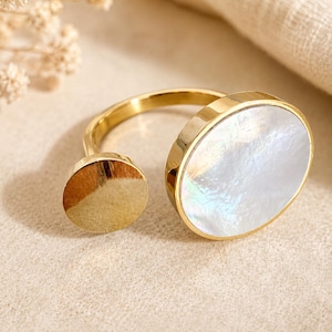Könnte beinhalten: Verstellbarer Ring in Goldoptik mit einem ovalen, schillernden Perlmuttstein. Der Ring hat ein schlichtes Design mit einem kleinen, runden Akzent. Ein Schmuckstück.