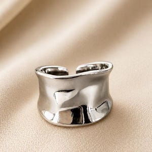 May include: A silver-colored ring with a textured, organic design. The ring has a wide band with an open design. The surface reflects light, creating highlights and shadows. The ring is likely made of metal and is a piece of jewelry.