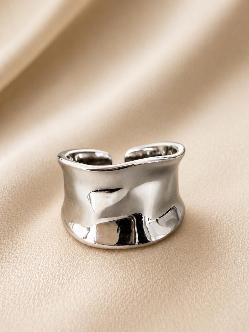 May include: A silver-colored ring with a textured, organic design. The ring has a wide band with an open design. The surface reflects light, creating highlights and shadows. The ring is likely made of metal and is a piece of jewelry.