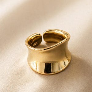 May include: A gold-colored ring with a wide, curved band. The ring has a smooth, polished surface and a slight opening at the top. The inside of the ring is stamped with "S925".