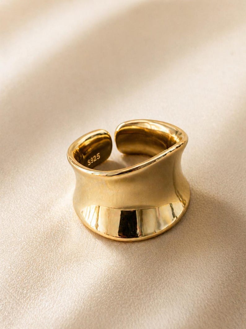 May include: A gold-colored ring with a wide, curved band. The ring has a smooth, polished surface and a slight opening at the top. The inside of the ring is stamped with "S925".
