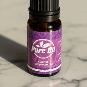 Könnte beinhalten: Kleine, braune Glasflasche von Pure Oil mit schwarzem Verschluss. Das Etikett ist lila mit weißem Text, der "Pure Oil" und "Lupines cold pressed oil" lautet. Die Flasche ist 100% natürlich.