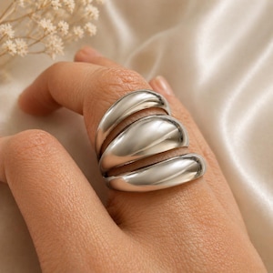 Könnte beinhalten: Ein silberfarbener Ring mit einem einzigartigen Design. Der Ring hat drei gestapelte, gebogene Bänder, die einen kühnen, modernen Look erzeugen. Die glatte, polierte Oberfläche reflektiert das Licht. Dieser Statement-Ring ist ein stilvolles Accessoire.