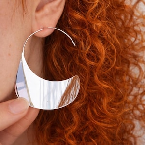 Könnte beinhalten: Ein großer, silberfarbener Creolen-Ohrring mit einem halbmondförmigen Design. Der Ohrring ist hochreflektierend und fängt das Licht ein, wodurch die Umgebung reflektiert wird. Der Ohrring wird von einer Person mit roten Locken getragen.