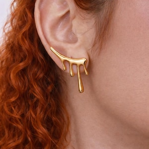 Könnte beinhalten: Ein goldfarbener Ohrschmuck mit tropfendem Flüssigkeitsdesign. Der Ohrring verläuft entlang des Ohrs und weist drei herabhängende Flüssigkeitstropfen auf. Der Schmuck wird am Ohr einer Person getragen.