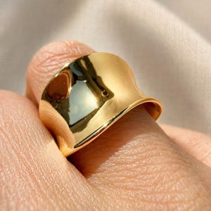 May include: A close-up of a gold-colored ring with a smooth, reflective surface. The ring has a unique, curved design, and it is worn on a finger. The ring's surface reflects the surrounding light.