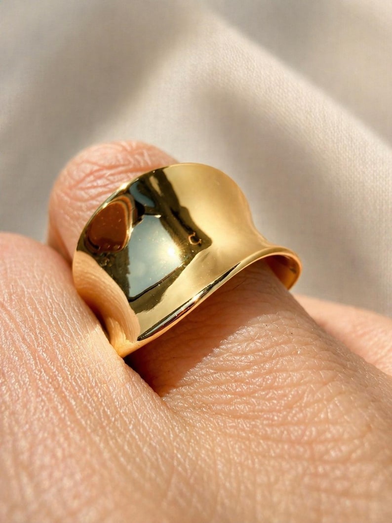 May include: A close-up of a gold-colored ring with a smooth, reflective surface. The ring has a unique, curved design, and it is worn on a finger. The ring's surface reflects the surrounding light.