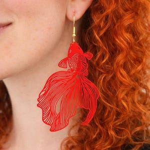 Könnte beinhalten: Roter Goldfisch-Ohrring mit aufwendigen Ausschnittdetails. Der Ohrring hängt an einem goldfarbenen Haken. Das Fischdesign hat fließende Flossen und einen detaillierten Körper, was ein auffälliges Accessoire ergibt.