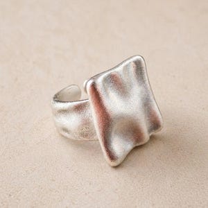 Könnte beinhalten: Ein silberfarbener Ring mit einem strukturierten, rechteckigen Mittelstück. Der Ring hat ein verstellbares Band und ein mattes Finish, was eine moderne, minimalistische Ästhetik schafft. Das Design weist eine fließende, organische Form auf.