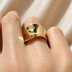 May include: A close-up shot of a gold-colored ring. The ring has a wide, curved band with a polished, reflective surface. The ring is on a finger, with the skin tone visible.
