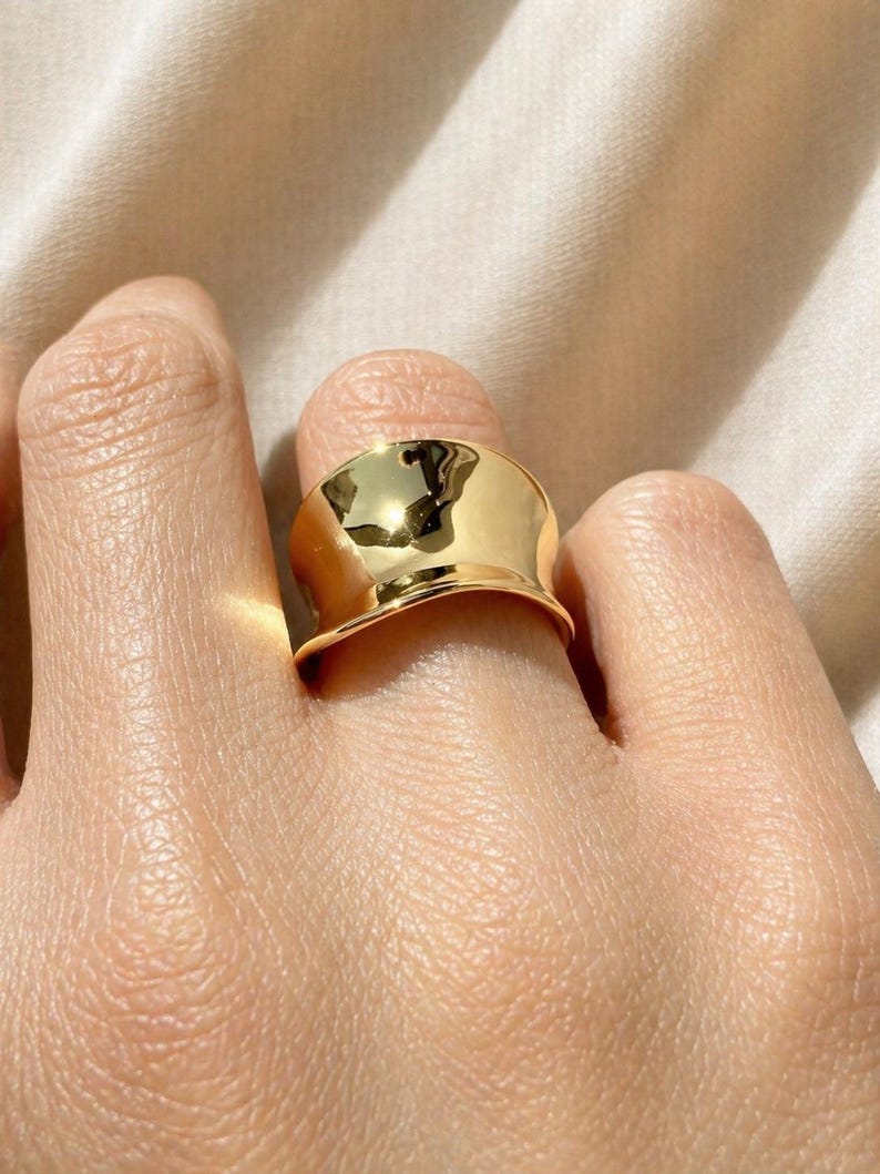 May include: A close-up shot of a gold-colored ring. The ring has a wide, curved band with a polished, reflective surface. The ring is on a finger, with the skin tone visible.