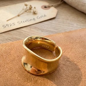 May include: A gold-colored, S925 sterling silver ring with a curved design. The ring is displayed on a brown surface, with a tag that reads "S925 Sterling Gold". The ring has a smooth, polished finish and a modern aesthetic.