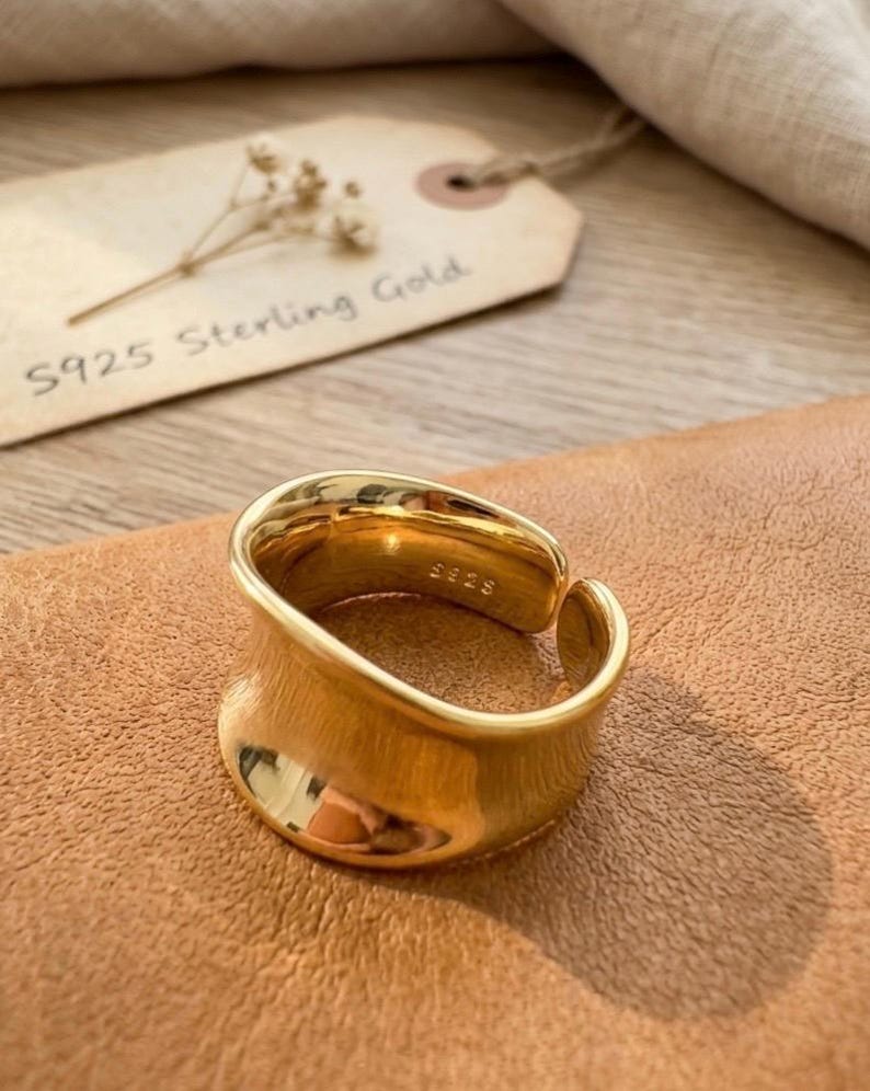 May include: A gold-colored, S925 sterling silver ring with a curved design. The ring is displayed on a brown surface, with a tag that reads "S925 Sterling Gold". The ring has a smooth, polished finish and a modern aesthetic.