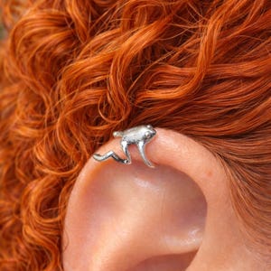 Puede incluir: Un pendiente de oreja de rana plateada. La rana está detallada con las patas extendidas, pareciendo trepar. El pendiente de oreja se lleva en la oreja de una persona, sobre un fondo de pelo rojo rizado y vibrante.