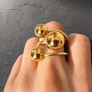 Könnte beinhalten: Ein goldfarbener Ring mit drei polierten, kugelförmigen Perlen unterschiedlicher Größe, die in einer Gruppe angeordnet sind. Die Ringschiene ist dünn und rund und trägt die drei glatten, reflektierenden Kugeln, die eine dreidimensionale Optik erzeugen.