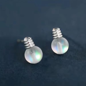 Könnte beinhalten: Ein Paar silberfarbene Ohrstecker in Form von Miniatur-Glühbirnen. Jede Glühbirne hat einen klaren, irisierenden Glaskörper und einen detaillierten silbernen Schraubsockel. Die Ohrringe sind vor einem dunkelblauen Hintergrund abgebildet.