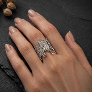 Könnte beinhalten: Ein silberner Ring mit einem unregelmäßigen, texturierten Design, das an geschmolzenes Metall oder tropfende Flüssigkeit erinnert. Die Ringschiene hat eine breite, offene Struktur mit vertikalen, unebenen Vorsprüngen.