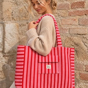 Puede incluir: Un bolso tote rectangular grande con rayas verticales en tonos alternos de rosa brillante y rojo. El bolso incluye un bolsillo frontal con solapa y una pequeña etiqueta rectangular blanca con el texto negro Bisou Bisou centrado.