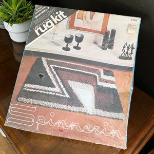 May include: A latch hook rug kit with a geometric design in black, white, and brown. The kit is labeled "geometric 7" and includes the text "latch hook rug kit" and "spinnerin".