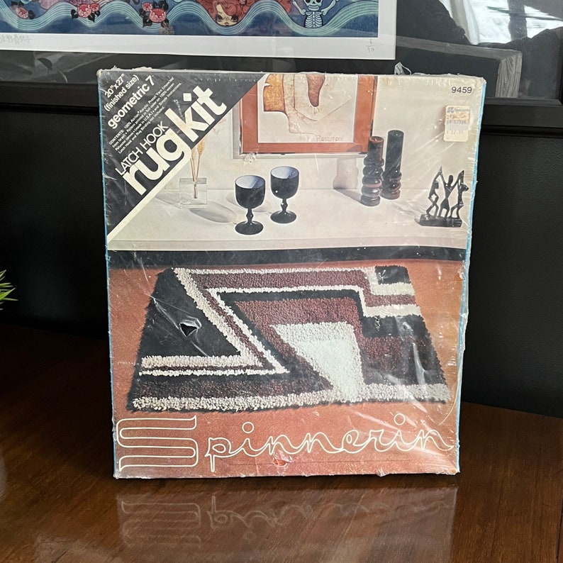 May include: A latch hook rug kit featuring a geometric design in shades of brown, white, and black. The kit includes yarn, instructions, and a latch hook tool. The finished rug size is 20" x 27".