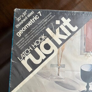 May include: A close-up of a latch hook rug kit packaging. The packaging is black and white with the words "geometric 7" and "latch hook rug kit" printed on it. The kit includes 100% acrylic yarn, a pattern printed on woven polyester canvas, and simple instructions. The finished rug size is 20 inches by 27 inches.