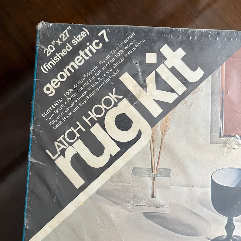 May include: A close-up of a latch hook rug kit packaging. The packaging is black and white with the words "geometric 7" and "latch hook rug kit" printed on it. The kit includes 100% acrylic yarn, a pattern printed on woven polyester canvas, and simple instructions. The finished rug size is 20 inches by 27 inches.