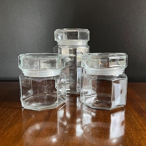 May include: Three clear glass canisters with octagonal lids. The canisters are of different sizes and are arranged in a row on a wooden surface.