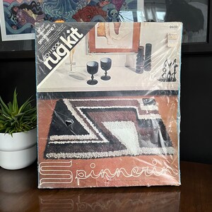 May include: A rug hooking kit with a geometric design. The kit includes yarn, a canvas, and instructions. The rug is black, white, and brown. The kit is labeled "Latch Hook Rug Kit" and "Geometric 7".
