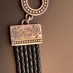 Op de afbeelding: Een zilverkleurige armbandsluiting met een decoratieve cirkelvormige bovenkant en een rechthoekige onderkant. De onderste sluiting is verbonden met verschillende strengen gevlochten zwart leer. De sluiting heeft een hart- en wervelontwerp.