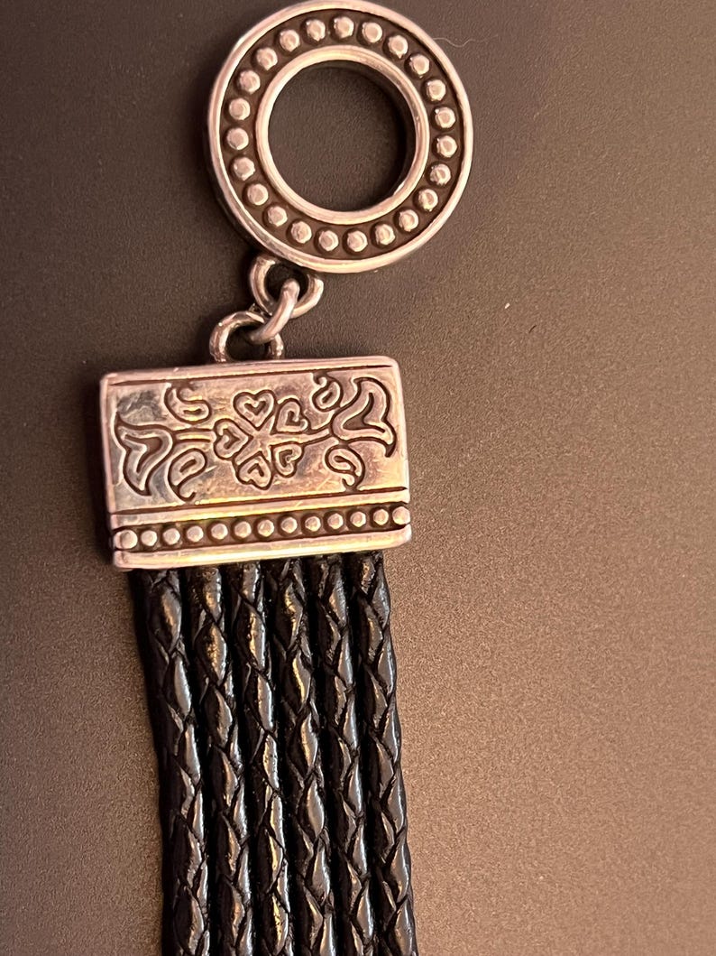 Op de afbeelding: Een zilverkleurige armbandsluiting met een decoratieve cirkelvormige bovenkant en een rechthoekige onderkant. De onderste sluiting is verbonden met verschillende strengen gevlochten zwart leer. De sluiting heeft een hart- en wervelontwerp.