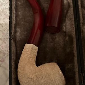 以下が含まれることがあります： 茶色の革ケースに入った、白と茶色のパイプ。赤いステムが付いている。